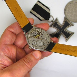 May include: A vintage wristwatch with a black leather strap, showcasing the intricate internal mechanisms. The watch is held near a black and white ribbon and a black iron cross medal. The strap is marked "Modena" and "Genuine Leather".