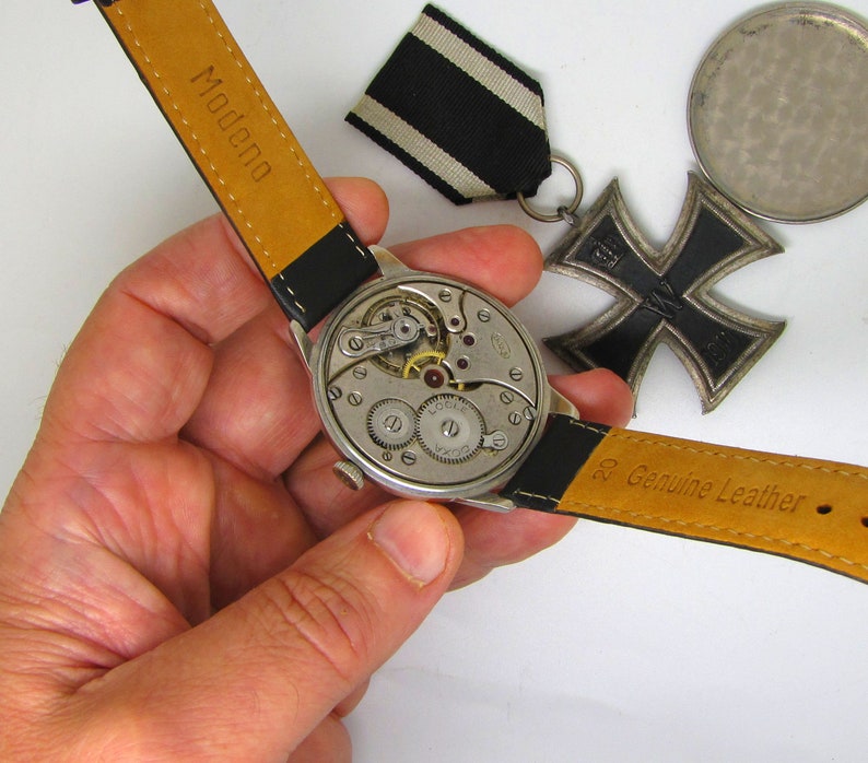 May include: A vintage wristwatch with a black leather strap, showcasing the intricate internal mechanisms. The watch is held near a black and white ribbon and a black iron cross medal. The strap is marked "Modena" and "Genuine Leather".