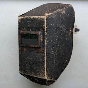 Peut inclure: Un casque de soudage noir vintage avec une fenêtre de visualisation rectangulaire. Le casque a une surface texturée et montre des signes d'usure, avec une lentille vert foncé et un cadre en métal rouillé. Un petit bouton noir est visible sur le côté.