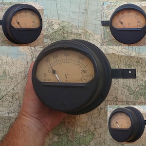 Puede incluir: Cuatro voltímetros analógicos vintage con cajas negras y caras blancas. Los voltímetros tienen una aguja que apunta a la lectura de voltaje. Los voltímetros están etiquetados con "V" para voltios. Los voltímetros están en varias posiciones, algunos están montados en una pared y otros se sostienen en una mano.