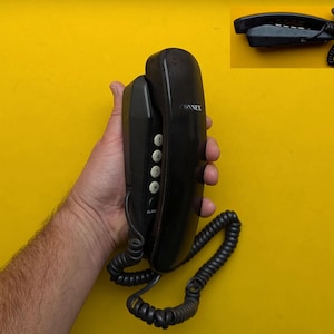 Könnte beinhalten: Schwarzes Connex-Telefon mit Spiralkabel vor gelbem Hintergrund. Das Telefon hat einen Hörer und eine Basis mit nummerierten Tasten. Eine Einlage zeigt den Hörer.