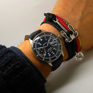 May include: A black-faced wristwatch with white numerals and a black leather band. The watch is next to a black and multi-colored bracelet with a silver clasp and a small silver charm.