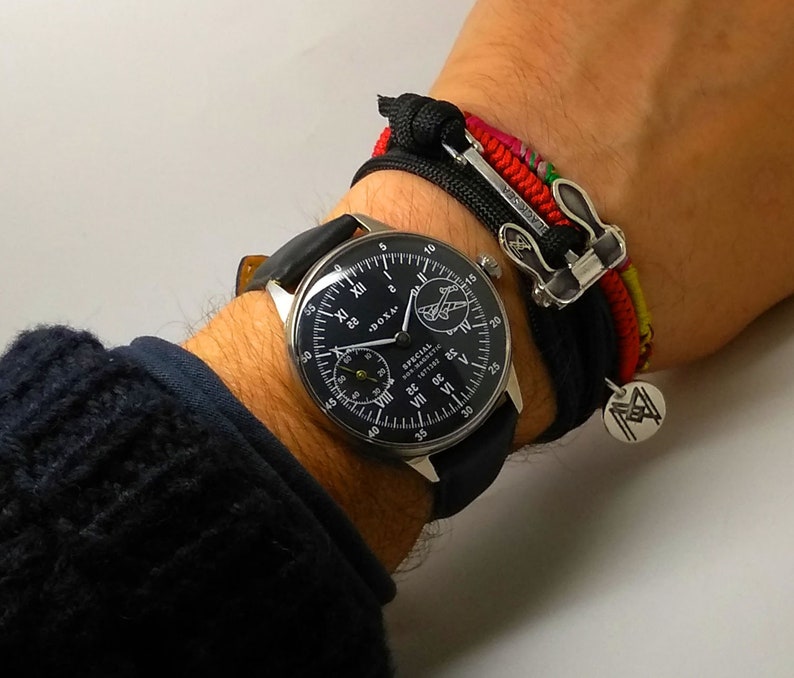 May include: A black-faced wristwatch with white numerals and a black leather band. The watch is next to a black and multi-colored bracelet with a silver clasp and a small silver charm.