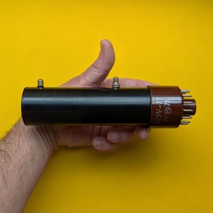 Pode incluir: Componente eletrónico cilíndrico preto e castanho com pinos, sobre um fundo amarelo. O componente tem duas pequenas protuberâncias prateadas na secção preta e texto na extremidade castanha. É uma válvula.
