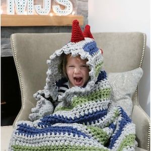 May include: A child wearing a gray, blue, and green striped crocheted blanket with a red and white hooded dinosaur head. The child is sitting in a chair and smiling.