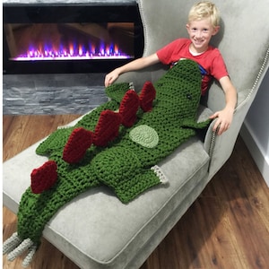 May include: A child sits on a grey armchair with a green and red crocheted dinosaur blanket. The blanket is shaped like a dinosaur with a long tail and a spiky back. The child is smiling and looking at the camera. #ItsAllInTheHookDesigns