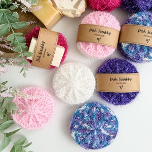 May include: A collection of handmade dish scrubbies in various colors, including pink, purple, and blue, with a white background. Some scrubbies have a brown paper band that reads "Dish Scrubby Handmade" with a heart symbol. Also shown are bars of soap and yarn.
