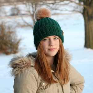 May include: A green knit beanie with a faux fur pom-pom. The hat is a deep forest green color and has a ribbed knit design. A small brown tag is attached to the side of the hat. The beanie is worn by a person wearing a tan jacket with a fur-lined hood.