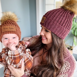 May include: A woman and a baby wearing matching brown knit hats with faux fur pom poms. The hats have personalized leather patches with the names "Rose Lynn" and "Mila". The woman is wearing a pink sweater and the baby is wearing a plaid shirt.