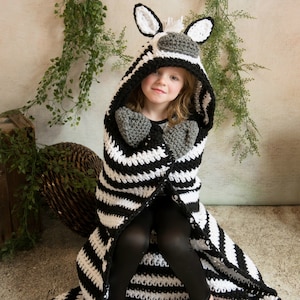 Könnte beinhalten: Eine gestrickte Zebra-Decke in Schwarz-Weiß mit Kapuze und angenähten Pfoten. Die Decke wird von einem Kind getragen, das auf einem grauen Teppich sitzt.