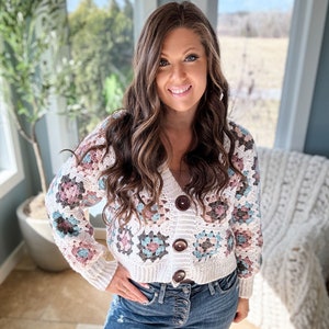 Op de afbeelding: Een wit gebreid vest met een granny square patroon in tinten roze, blauw en grijs. Het vest heeft een V-hals en bruine knopen. Het model draagt het vest met een blauwe jeans.