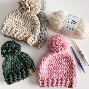 May include: Three crocheted hats with pom-poms in different colors: beige with grey, dark green, and pink. The hats are on a white surface with a ball of yarn and a crochet hook.
