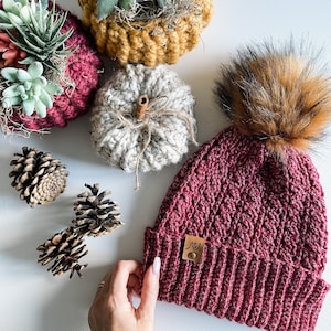 Puede incluir: Un gorro de punto burdeos con un pompón de piel sintética. El gorro tiene una etiqueta de cuero marrón con la palabra "Mels" escrita en ella. El gorro está rodeado de decoraciones de otoño, incluyendo calabazas de punto y plantas suculentas.