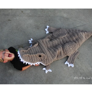 May include: A child is lying in a crocheted dinosaur blanket. The blanket is brown with white accents. The child is wearing glasses and has their mouth open in a scream.