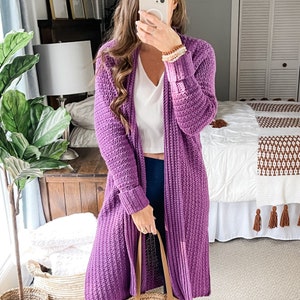 May include: A long, purple cardigan with a ribbed texture. The cardigan is open at the front and has long sleeves with ribbed cuffs. The person is wearing a white vest top and blue jeans.