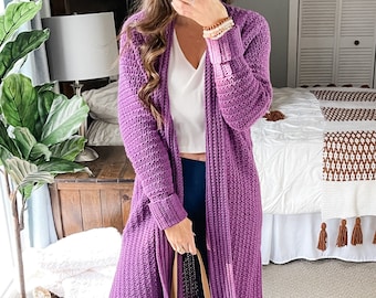 Puede incluir: Un cárdigan largo de color morado con textura de canalé. El cárdigan está abierto por delante y tiene mangas largas con puños de canalé. La persona lleva un top blanco sin mangas y unos vaqueros azules.