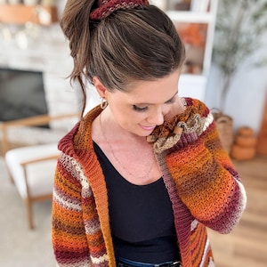 May include: A hooded, long-sleeved, crocheted cardigan in a warm color palette of orange, burgundy, cream, and brown stripes. The woman in the image is wearing a black tank top and blue jeans. A matching crocheted scrunchie is in her hair.