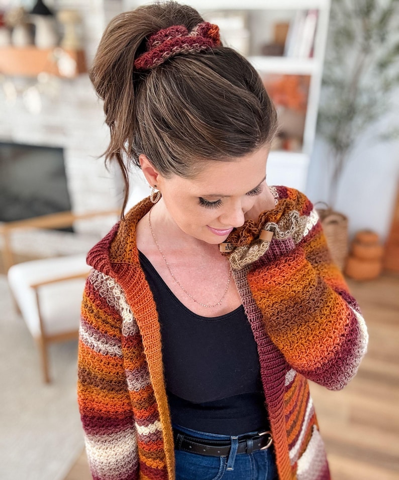 May include: A hooded, long-sleeved, crocheted cardigan in a warm color palette of orange, burgundy, cream, and brown stripes. The woman in the image is wearing a black tank top and blue jeans. A matching crocheted scrunchie is in her hair.