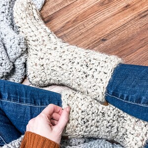 May include: A pair of crocheted cream-coloured socks with a textured stitch pattern. The socks are worn by a person sitting on a wooden floor with a grey blanket.