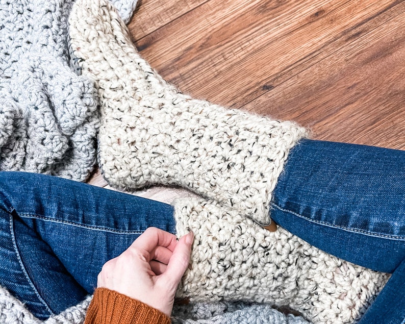 May include: A pair of crocheted cream-coloured socks with a textured stitch pattern. The socks are worn by a person sitting on a wooden floor with a grey blanket.