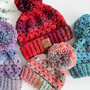 May include: Three crocheted hats with pom-poms in various colors, including red, pink, blue, and purple. Each hat has a small leather tag with the text "Mel's Stitch Designs".