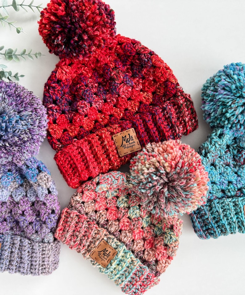 May include: Three crocheted hats with pom-poms in various colors, including red, pink, blue, and purple. Each hat has a small leather tag with the text "Mel's Stitch Designs".