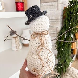 May include: A white crocheted snowman with a black and white speckled top hat and a brown twine bow. The snowman is standing on a wooden surface.
