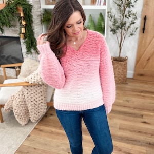 May include: A pink and white ombre crochet sweater with a V-neck and long sleeves. The sweater is paired with blue jeans. The sweater's color transitions from a deep pink at the top to white at the bottom.