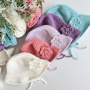 May include: A collection of handmade crochet hats in various pastel colors, including white, pink, purple, and blue. Each hat features a crocheted flower and ties. Yarn and floral accents are in the background.