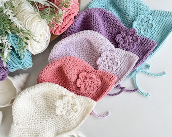 May include: A collection of handmade crochet hats in various pastel colors, including white, pink, purple, and blue. Each hat features a crocheted flower and ties. Yarn and floral accents are in the background.