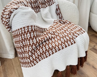 May include: A crochet throw blanket featuring a repeating geometric pattern in white and brown yarn. The blanket includes solid white horizontal bands and is finished with brown fringe tassels along the bottom edge of the fabric.
