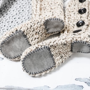 May include: A pair of beige crocheted slipper boots with suede soles. The boots have a button detail on the top.