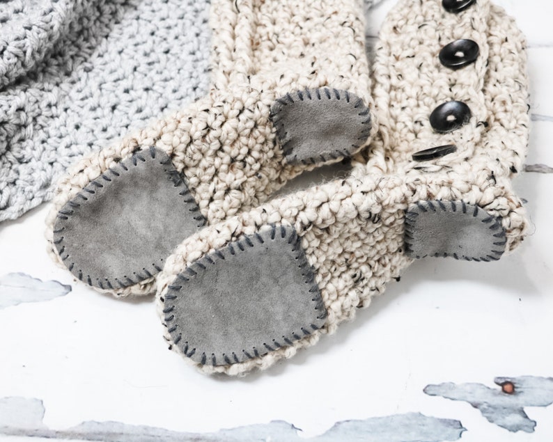 May include: A pair of beige crocheted slipper boots with suede soles. The boots have a button detail on the top.