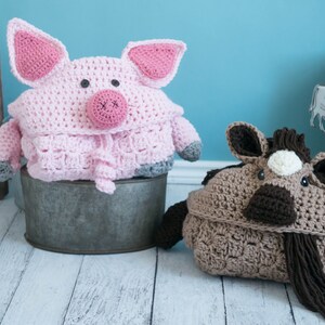 Pu&ograve; includere: Due coperte di peluche per animali in crochet, un maiale rosa e un cavallo marrone, sono seduti su una superficie di legno bianco. Il maiale &egrave; seduto in un secchio di metallo e il cavallo &egrave; sul pavimento.