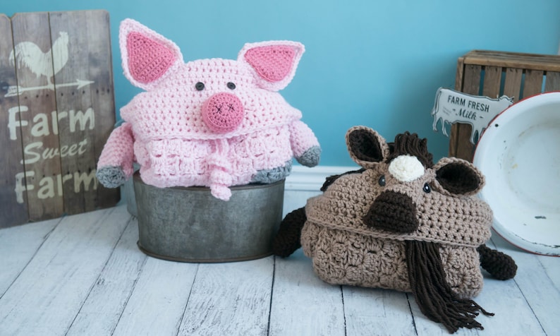 Pu&ograve; includere: Due coperte di peluche per animali in crochet, un maiale rosa e un cavallo marrone, sono seduti su una superficie di legno bianco. Il maiale &egrave; seduto in un secchio di metallo e il cavallo &egrave; sul pavimento.