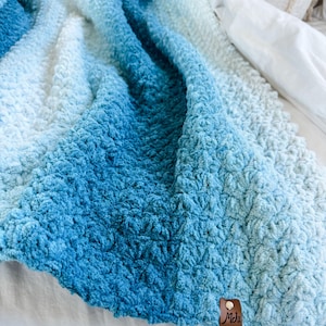 May include: A plush, textured blanket in shades of blue, transitioning from deep ocean hues to a soft, light blue. The blanket has a dense, raised pattern and a small, brown leather tag with the initials "Mds".