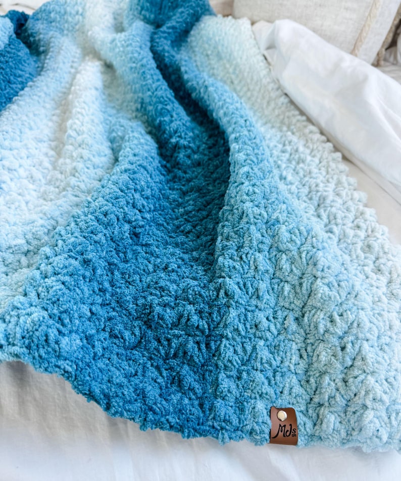 May include: A plush, textured blanket in shades of blue, transitioning from deep ocean hues to a soft, light blue. The blanket has a dense, raised pattern and a small, brown leather tag with the initials "Mds".