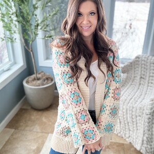 May include: A cream-coloured crochet cardigan with a granny square pattern in shades of pink, blue, and brown. The cardigan has long sleeves and a relaxed fit.