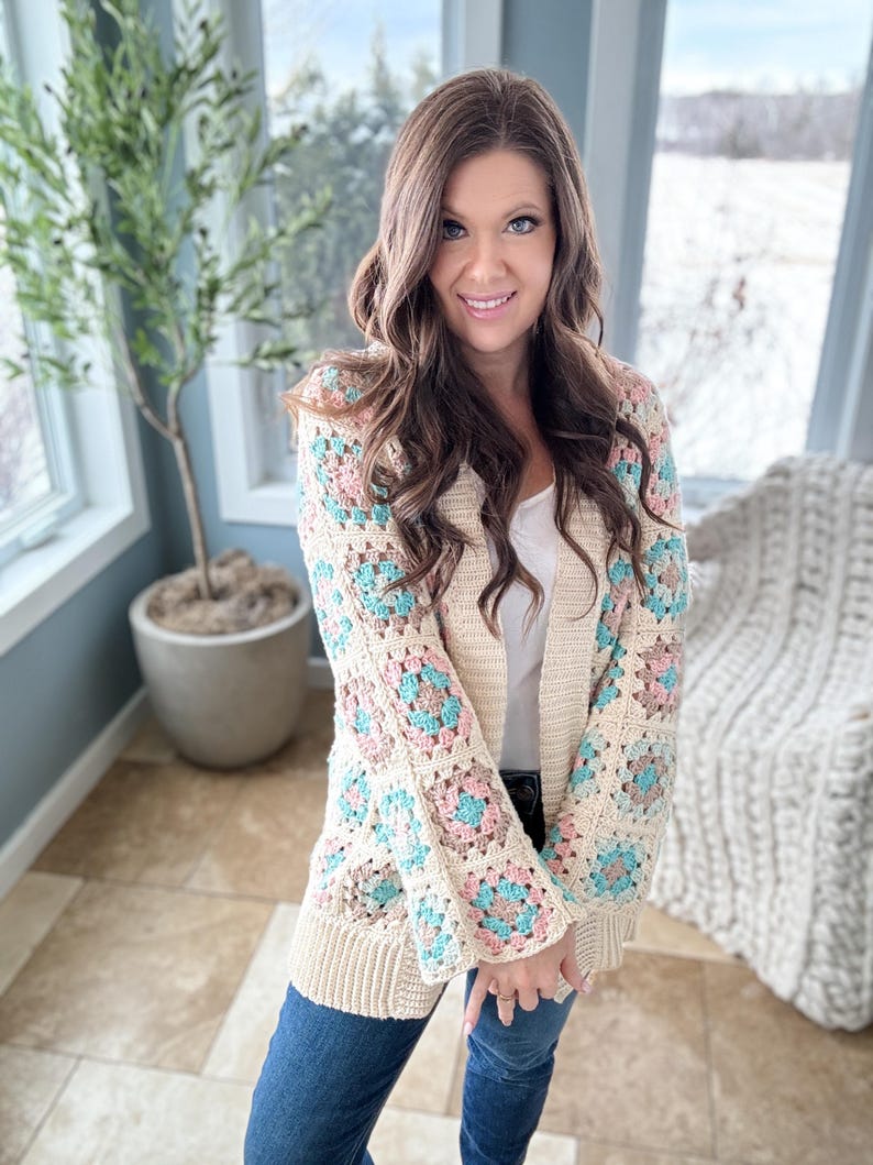 May include: A cream-coloured crochet cardigan with a granny square pattern in shades of pink, blue, and brown. The cardigan has long sleeves and a relaxed fit.