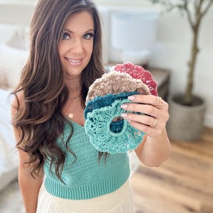 May include: A collection of handmade crochet scrunchies in various colors, including teal, brown, and red. The scrunchies are displayed against a soft background, showcasing their textured, detailed design. These hair accessories are perfect for adding a touch of handmade charm to any hairstyle.