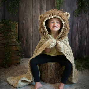 May include: A child wearing a crocheted lion costume with a brown hooded cape and matching mittens. The costume is made of beige yarn with brown accents. The child is sitting on a tree stump in front of a wooden background.