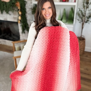 May include: A handmade, crochet blanket in a gradient of red and pink hues. The blanket is held up, showcasing the textured stitch pattern and the color transition from light pink to deep red. The blanket appears soft and cozy.