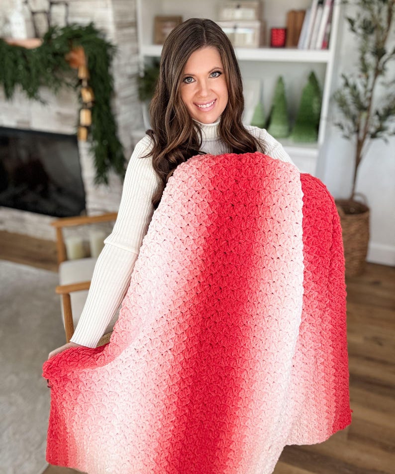 May include: A handmade, crochet blanket in a gradient of red and pink hues. The blanket is held up, showcasing the textured stitch pattern and the color transition from light pink to deep red. The blanket appears soft and cozy.