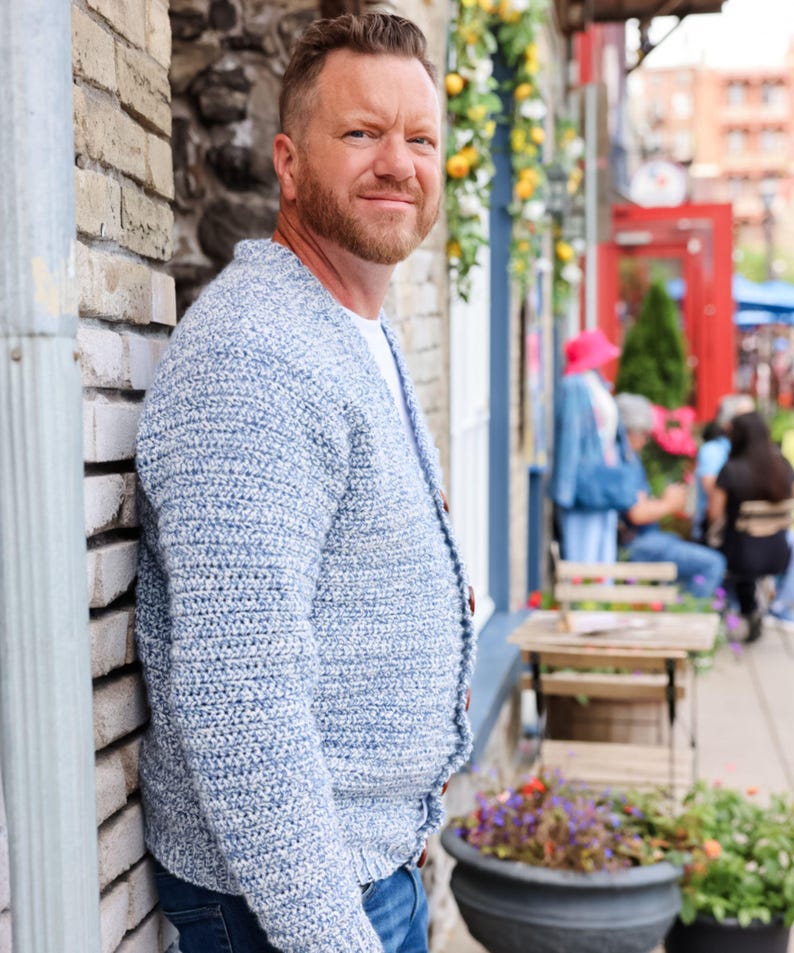 May include: A light blue crochet cardigan with wooden buttons. The open-front sweater has a textured knit pattern and is worn over a white t-shirt. The cardigan is a casual, comfortable style.