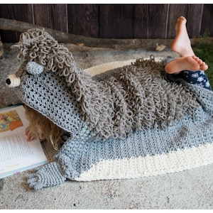 May include: A grey and white crocheted hedgehog blanket with a hood and mittens. The blanket is made with a fluffy yarn and has a cute hedgehog face on the hood.