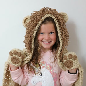 May include: A child wearing a brown and beige crocheted lion hooded scarf with paw-shaped hand warmers. The scarf has a fluffy mane and the child is smiling.