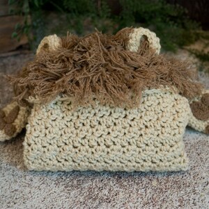 May include: A crocheted lion costume for a baby, featuring a tan body with brown fur accents. The costume has a hood with a lion's mane and two small paws.