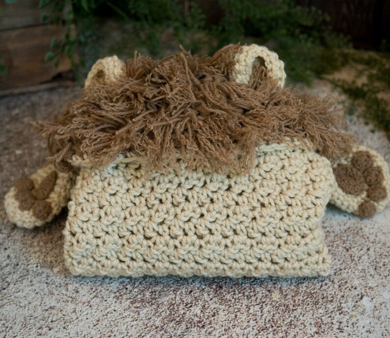 May include: A crocheted lion costume for a baby, featuring a tan body with brown fur accents. The costume has a hood with a lion's mane and two small paws.