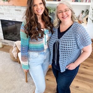 May include: Two women smiling, wearing handmade crochet cardigans. One cardigan is a mix of blue, green, and brown, the other is light blue. Both are paired with jeans. The background includes a fireplace and a bookshelf.
