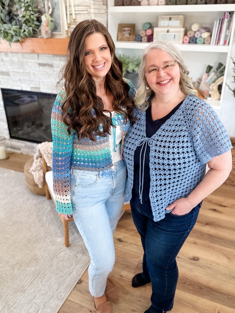 May include: Two women smiling, wearing handmade crochet cardigans. One cardigan is a mix of blue, green, and brown, the other is light blue. Both are paired with jeans. The background includes a fireplace and a bookshelf.